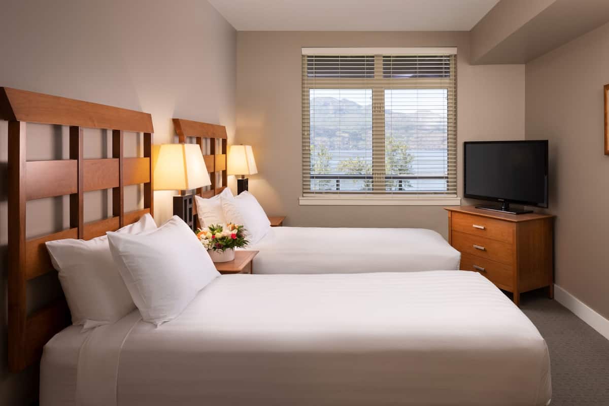 Una habitación de hotel con dos camas, televisor, cabecero de madera, mesitas de noche con lámparas y una ventana con persianas.
