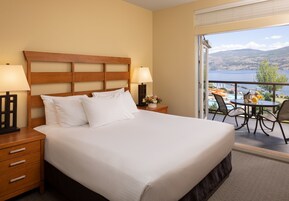 Habitación de hotel con una cama grande, dos mesitas de noche con lámparas, una pequeña mesa con un arreglo floral, y un balcón con mesa y sillas con vistas a un cuerpo de agua y colinas.
