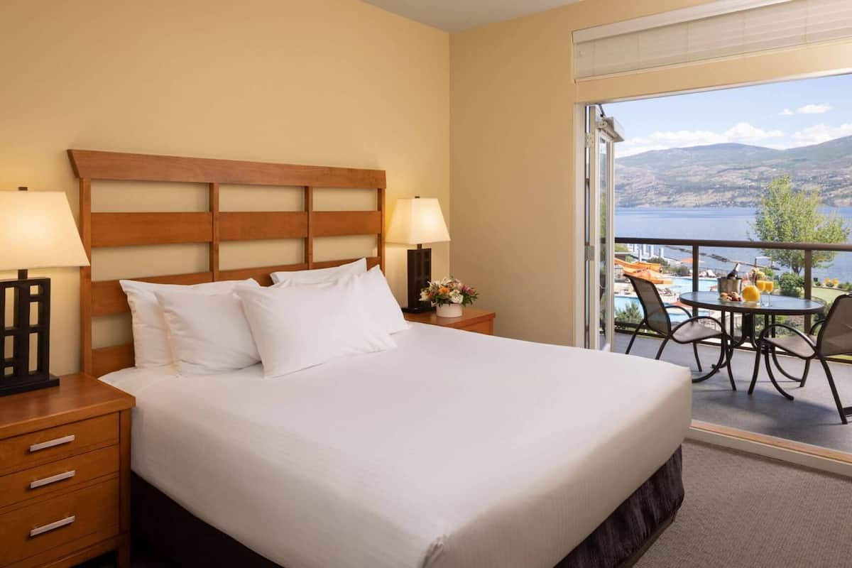 Habitación de hotel con una cama grande, dos lámparas de noche, un cabecero de madera, una mesita de noche con un florero y un balcón con mesa y sillas con vistas a un cuerpo de agua.