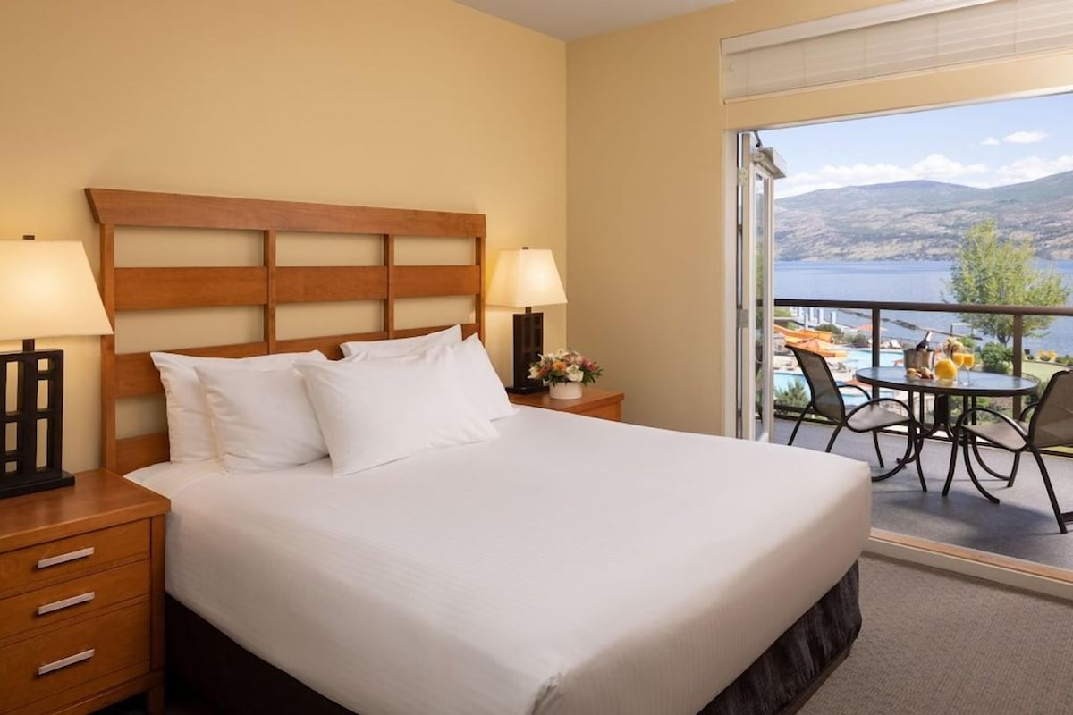 Habitación de hotel con una cama grande, dos lámparas de noche, una mesita de noche, una pequeña mesa con un florero y un balcón con mesa y sillas.