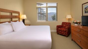 A hotel room with a large bed, a wooden headboard, a window with blinds, a red floral armchair, a wooden dresser, and a television.