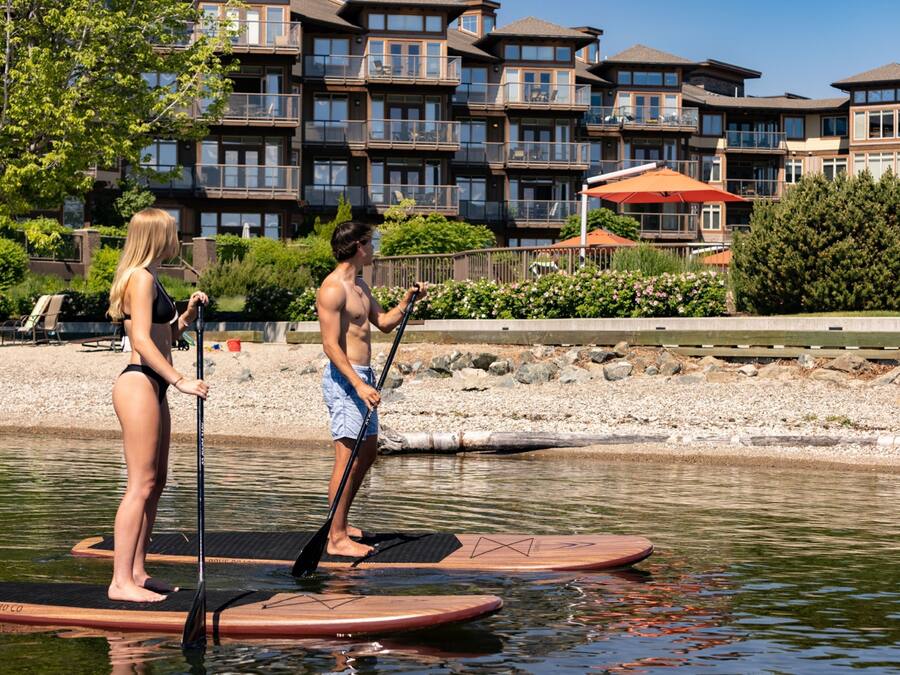 Deux personnes sur des planches à pagaie dans une étendue d’eau calme, avec un immeuble d’habitation en arrière-plan.
