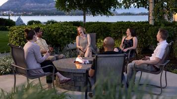 A group of people seated around a stone fire pit in an outdoor setting with a lake and hills in the background.
