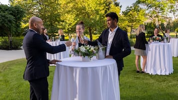 Ryhmä ihmisiä kohottaa maljan ulkoilmatapahtumassa. Pyöreät pöydät on katettu valkoisilla pöytäliinoilla, ja niiden ympärillä on vehreää kasvillisuutta.