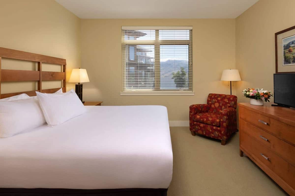 Una habitación de hotel con una cama grande, cabecera de madera, una ventana con persianas, un sillón rojo, un aparador de madera y un televisor.