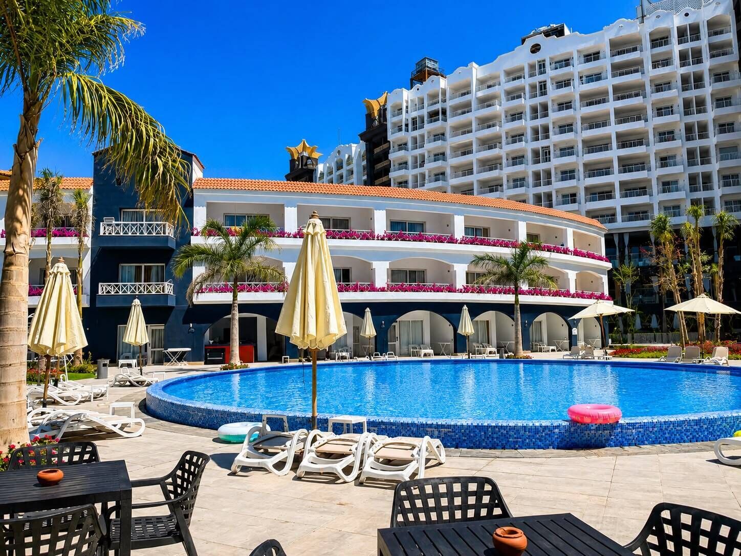 Zona de piscina de hotel con sillones, sombrillas y un edificio grande al fondo.