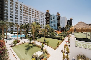 A hotel with multiple floors, a swimming pool, and a well-maintained garden with palm trees and a thatched-roof structure.