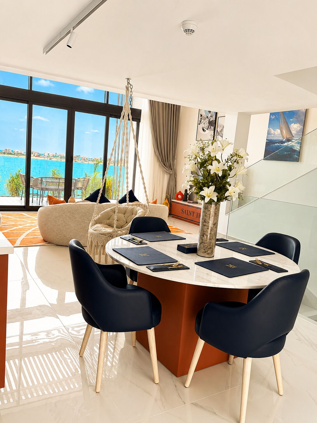 Een moderne eethoek met uitzicht op zee, een hangstoel en een vaas met bloemen.