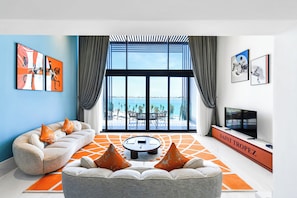 Una sala de estar moderna con un sofá, una mesa de centro y un televisor. Hay ventanas amplias con vistas al mar y a la ciudad.