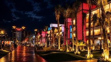 A nighttime view of a hotel with a paved walkway, palm trees, and illuminated buildings.