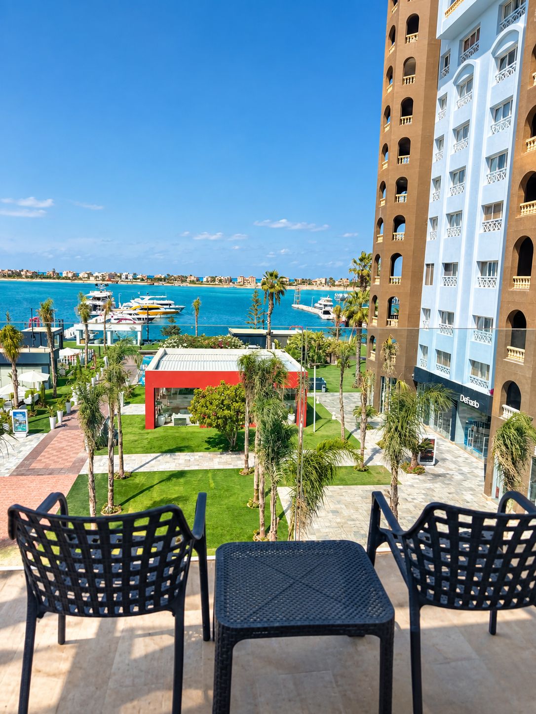 Ein Hotelbalkon mit Blick auf das Meer und Boote, ein rotes Gebäude und Palmen.