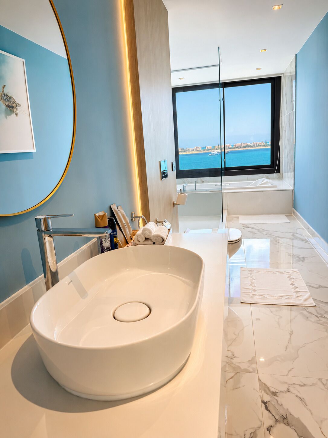 A modern bathroom with a large oval sink, a glass shower enclosure, and a view of the sea through a large window.