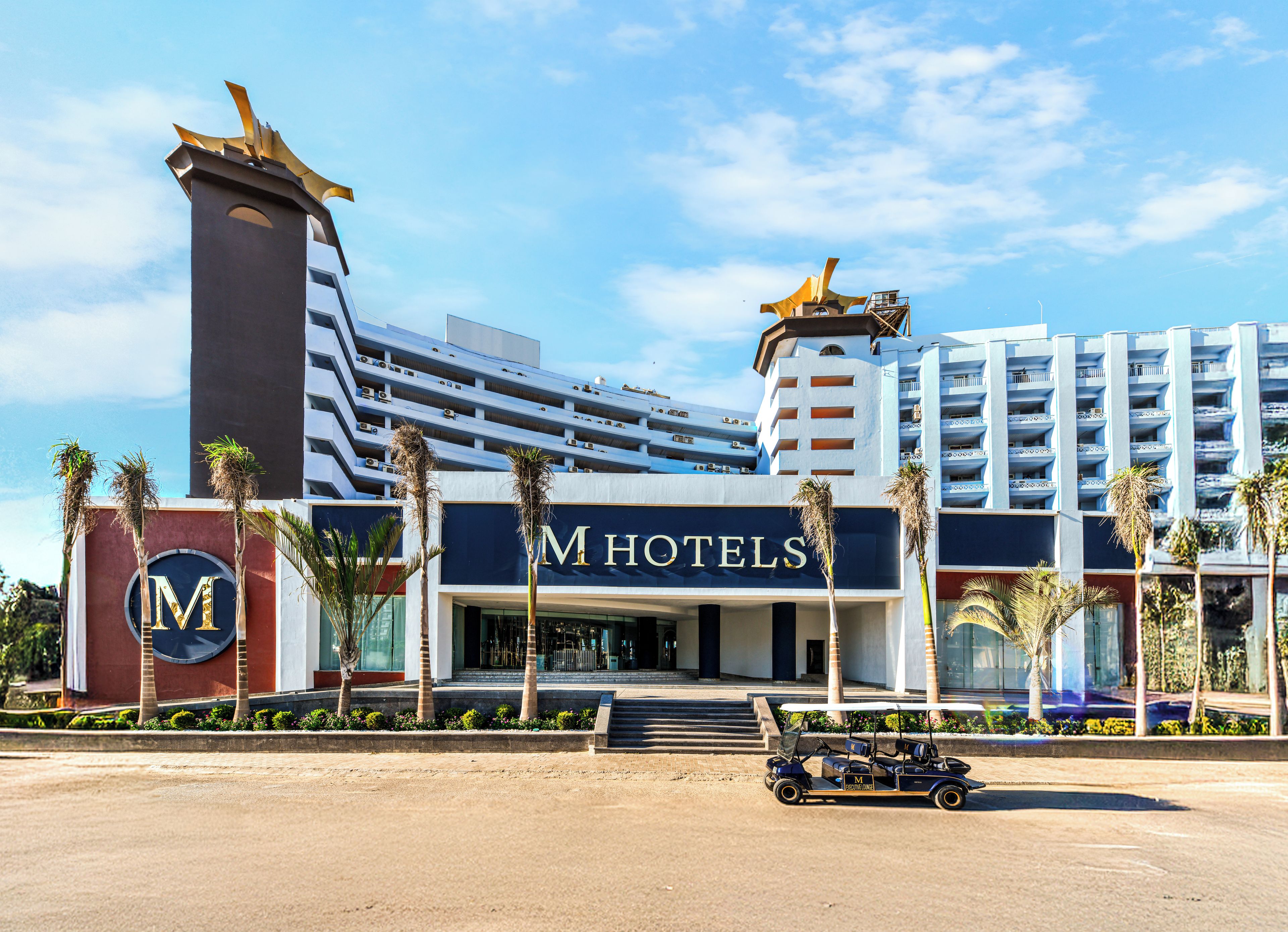 A hotel with a prominent 'M HOTELS' sign, a large vertical structure with a golden top, and a golf cart in the foreground.