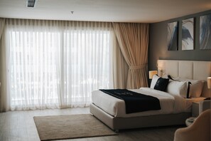 A modern hotel room with a large bed, a bedside table with lamps, a view through sheer curtains, and abstract art on the wall.