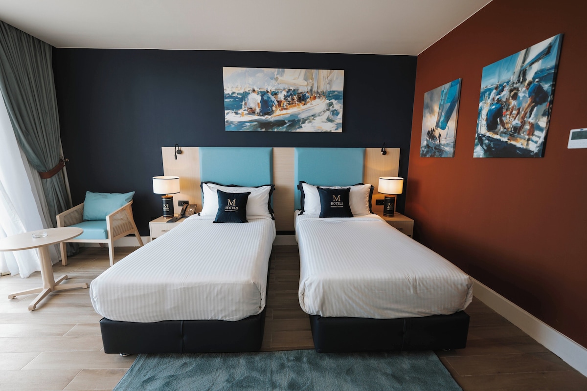 A hotel room with two beds, a small table, a chair, and artwork on the walls.