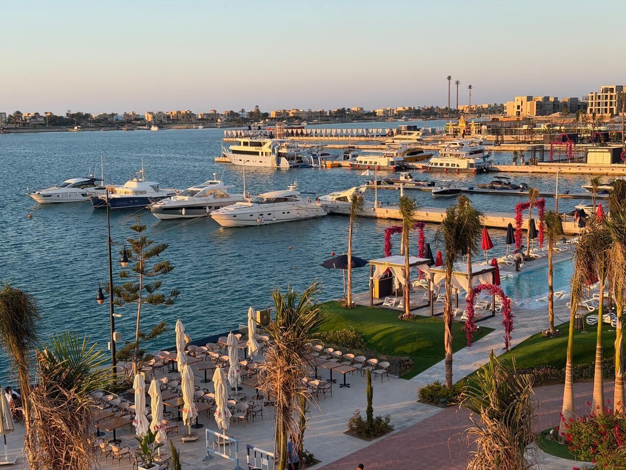A marina with boats docked, a pool area with umbrellas, and a cityscape in the background.