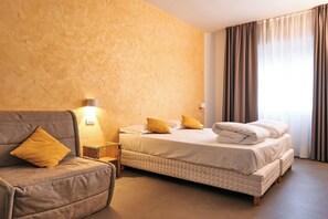 A hotel room with a sofa, a bed with pillows, a nightstand, and a wall-mounted lamp.
