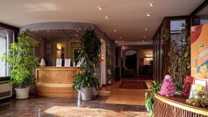 A hotel reception area with a curved wooden reception desk, potted plants, and a glass display case. - Due Ragni (Villorba)