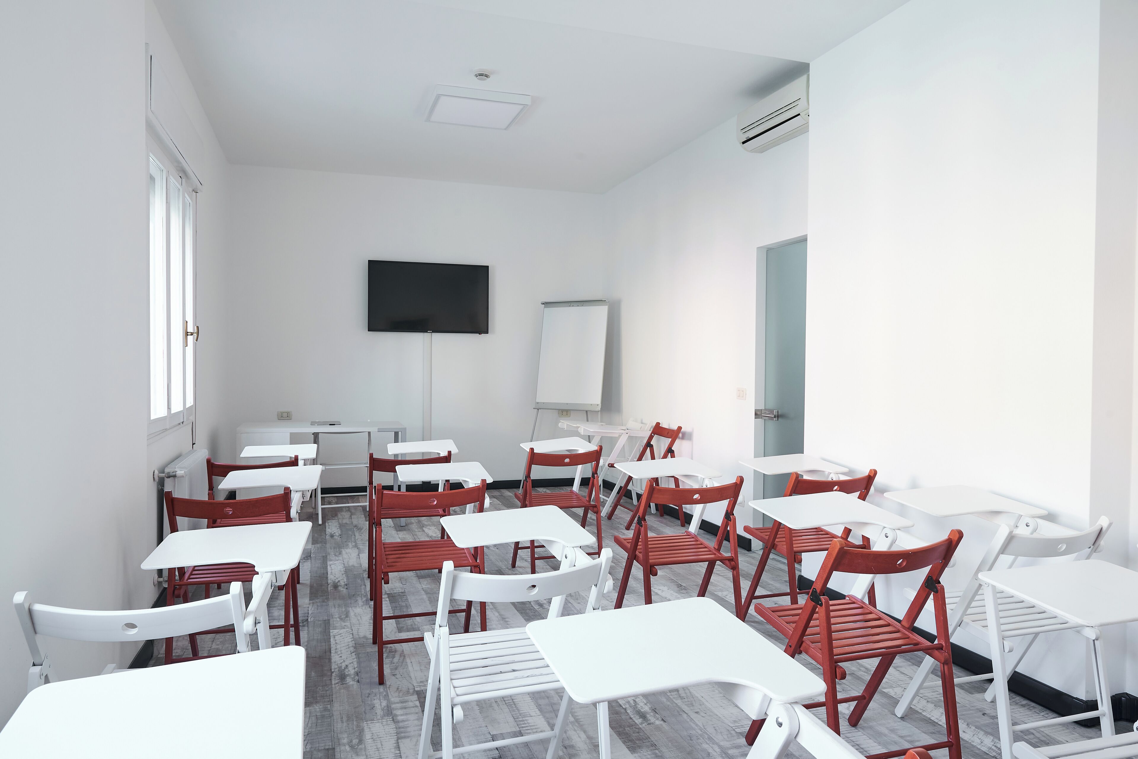 Une salle de classe avec des tables blanches et des chaises rouges, un tableau blanc et un téléviseur fixé au mur.