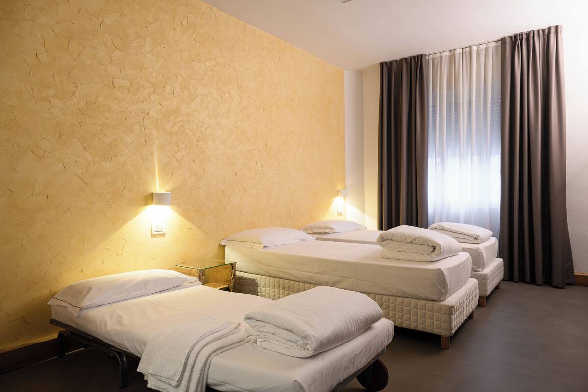 A hotel room with three beds, a textured beige wall, and a large window with curtains.