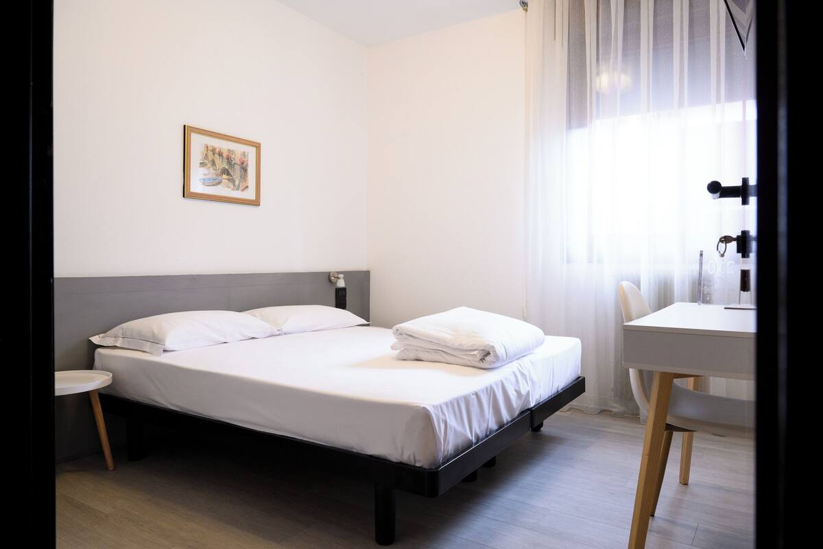 A neatly made bed with white linens, a small bedside table, and a chair at a desk with a lamp.