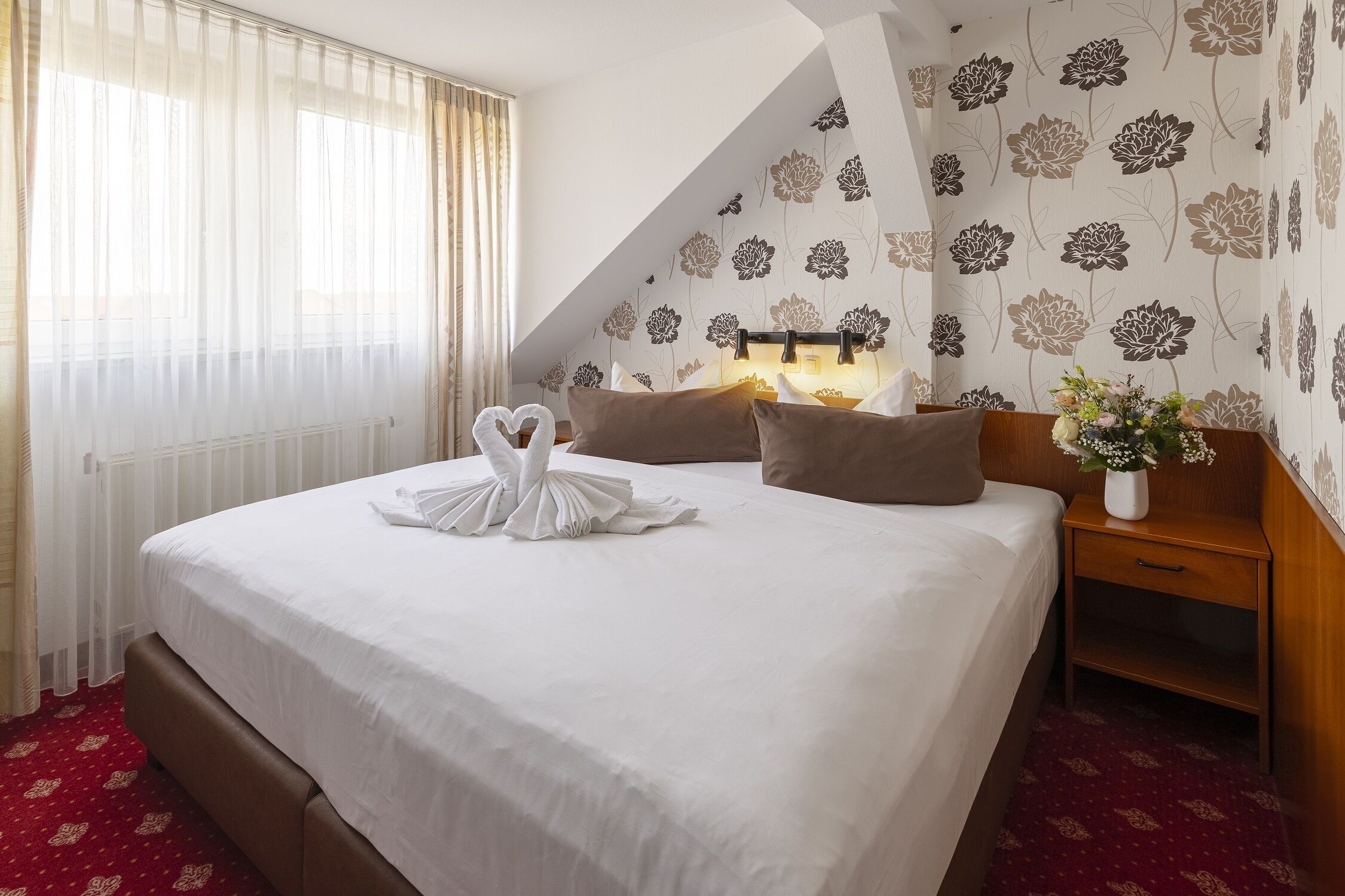 Habitació d'hotel amb un llit ben fet, una tauleta de nit amb un ram de flors i una paret amb motius florals.