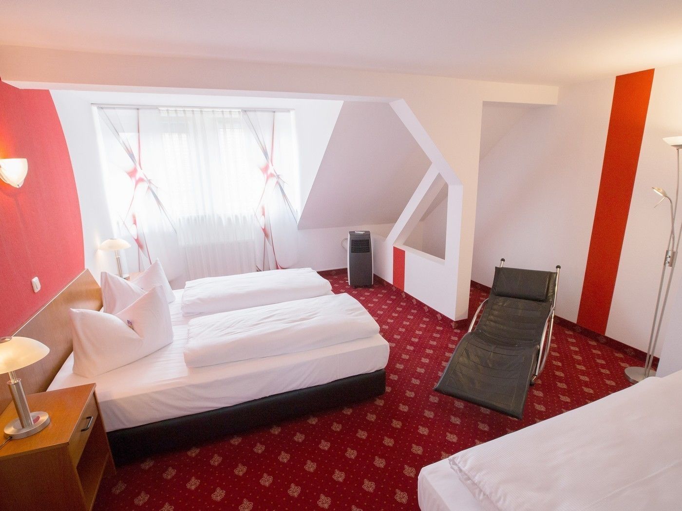 A hotel room with two beds, a red carpet, a standing chair, a heater, and a window with curtains.