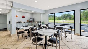 A modern dining area with multiple tables and chairs, a kitchen in the background, and large windows offering a view of the outdoors.