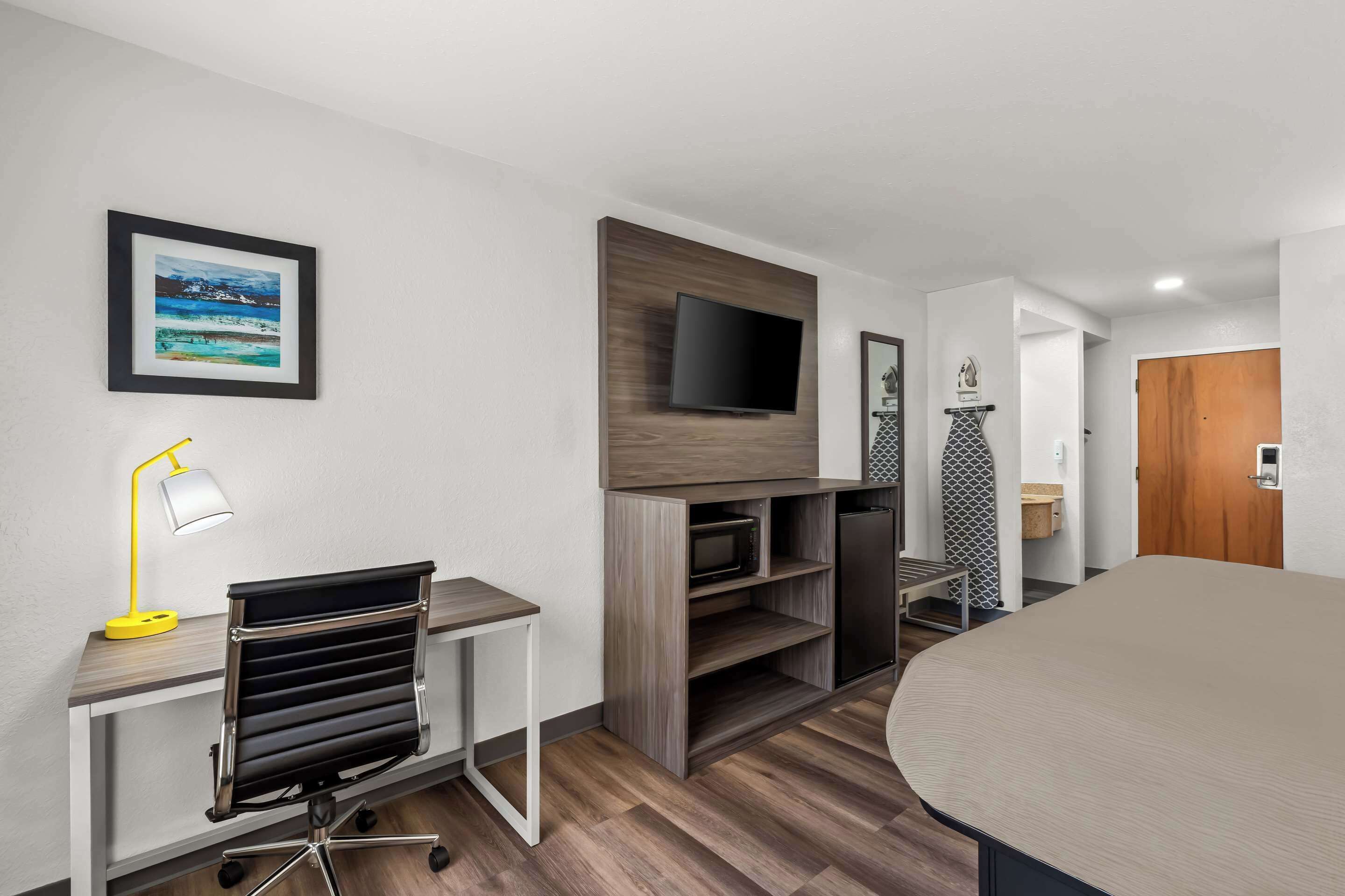 Habitación de hotel moderna con televisión de pantalla plana montada en un panel de madera, un escritorio con lámpara de mesa amarilla, una silla de oficina negra y una cama grande.