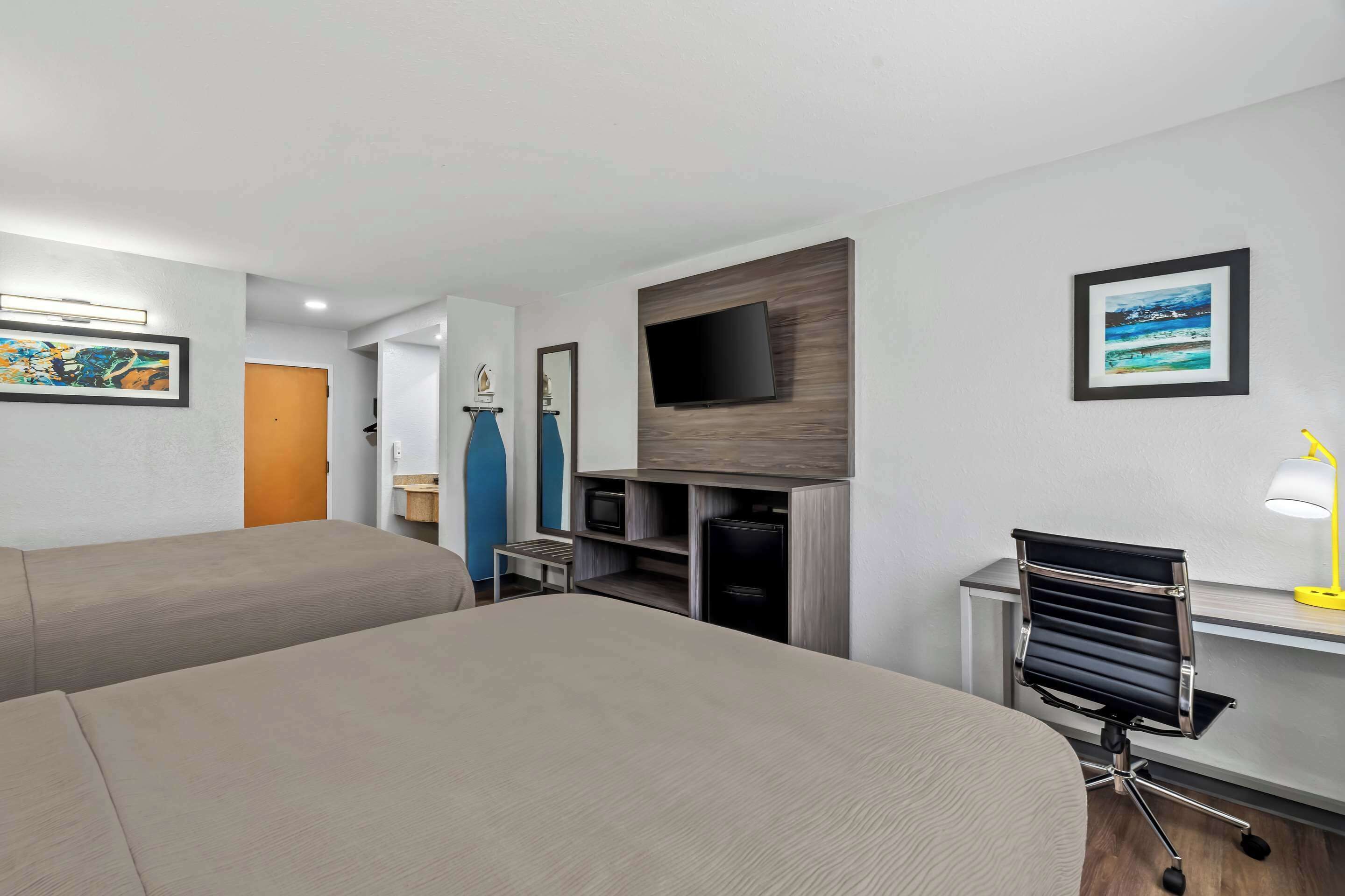 Ein Hotelzimmer mit zwei Betten, einem Schreibtisch mit Stuhl, einem an der Wand befestigten Fernseher und einem gerahmten Bild.