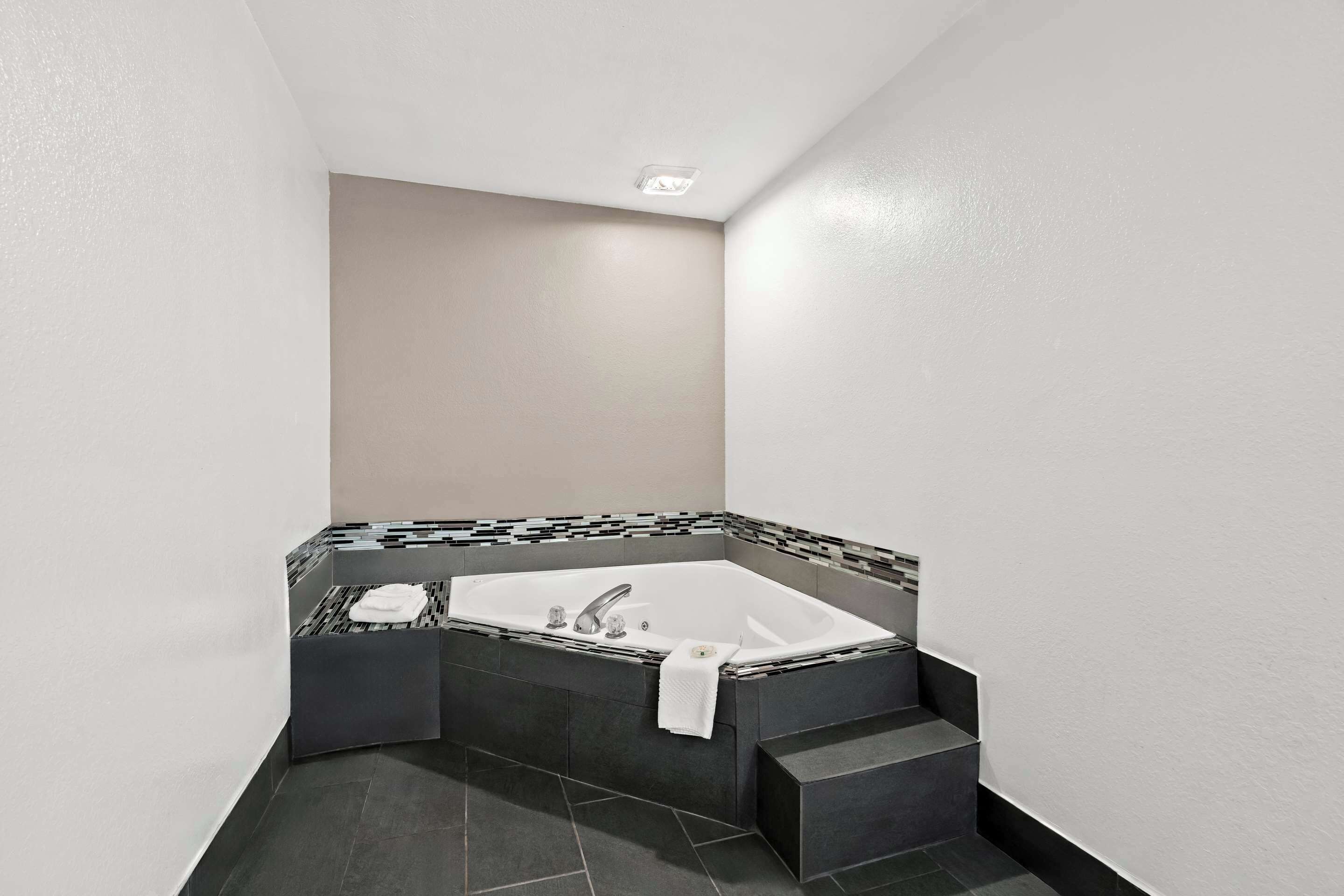 Une salle de bain moderne avec une baignoire d’angle, des carreaux foncés et une serviette blanche posée sur le bord.