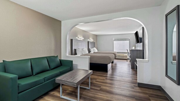 A modern hotel room with a green sofa, a wooden coffee table, a bed with a gray headboard, a desk with a computer, and a TV mounted on the wall.