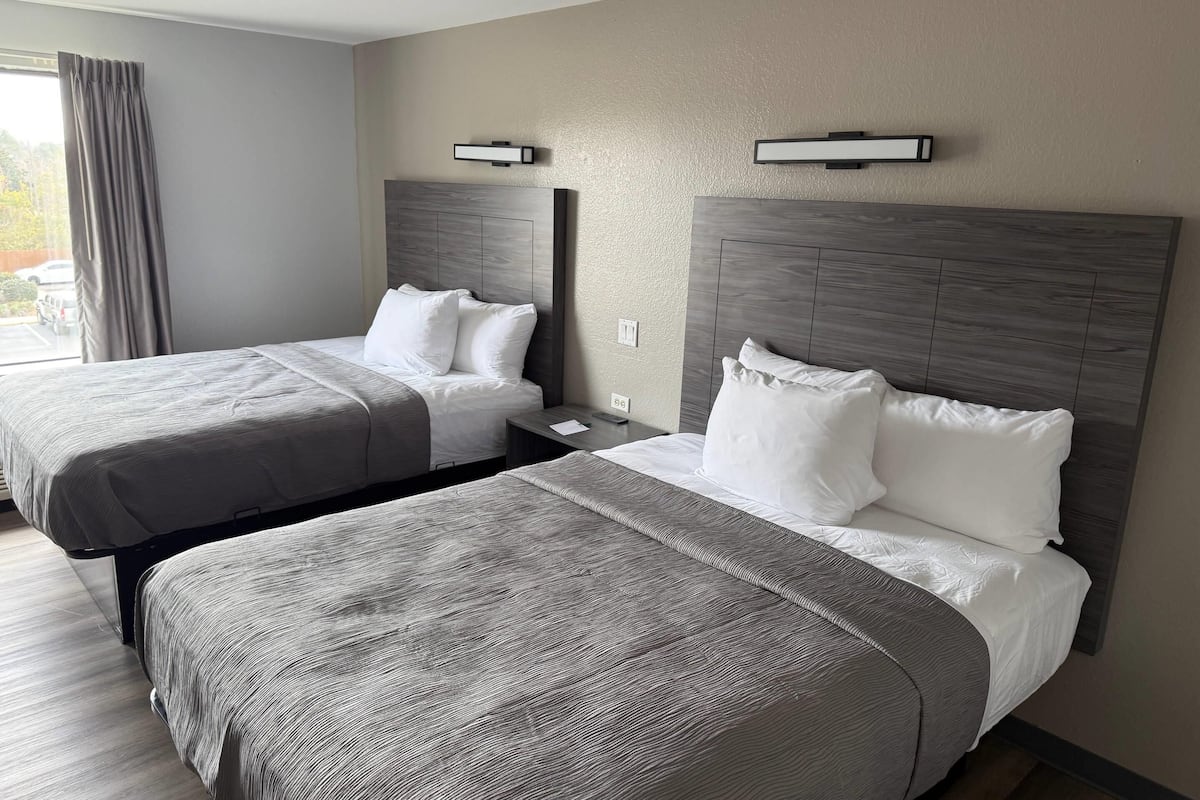 Ett hotellrum med två sängar, ett stort fönster med gardiner och en väggmonterad lampa.