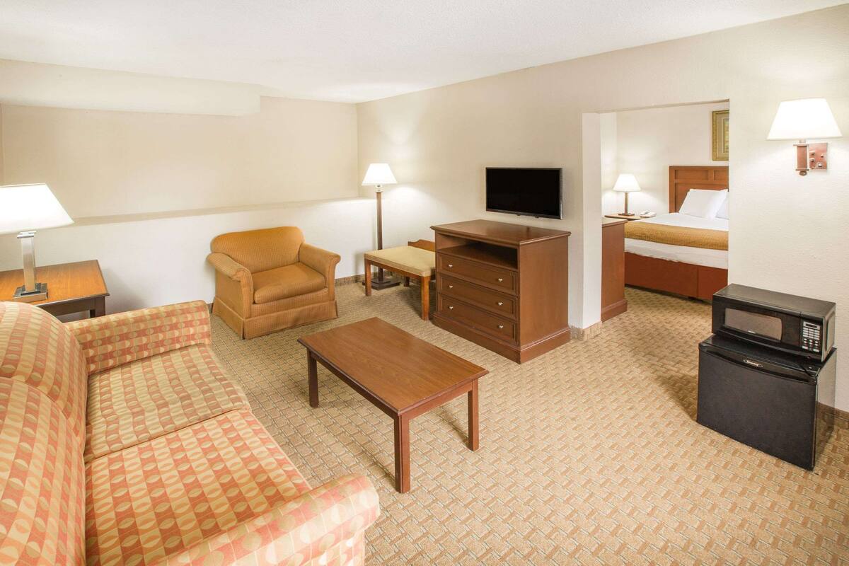 A hotel room with a sofa, a coffee table, a TV, a desk with a lamp, a bed, and a microwave.