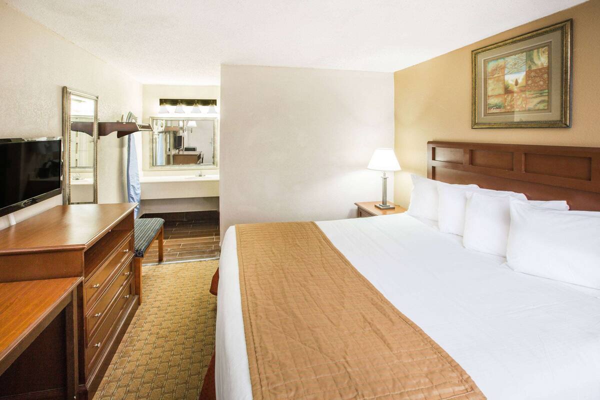 A hotel room with a large bed, a desk with drawers, a television, a mirror, a lamp, and a painting on the wall.