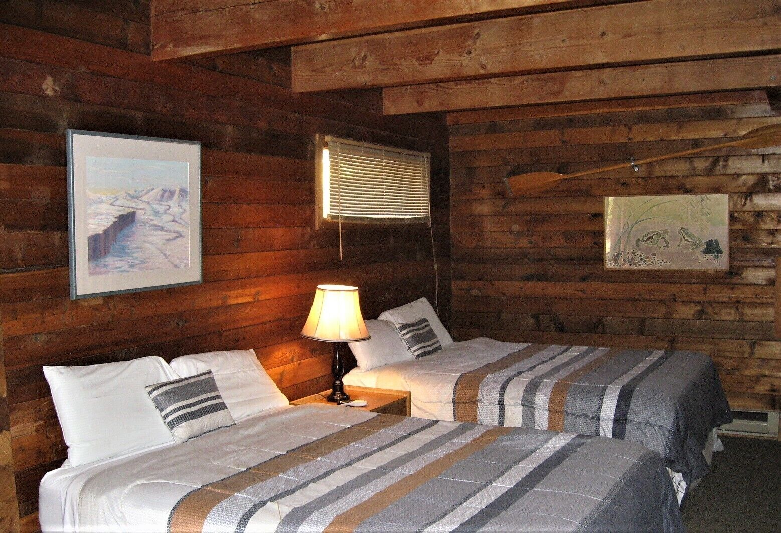 A wooden cabin room with two beds, a lamp, and framed pictures on the walls.