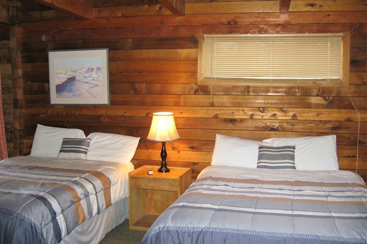 Acogedora habitación tipo cabaña con dos camas, cabecero de madera, un cuadro y una ventana con persianas.