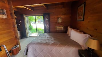 A wooden cabin room with a bed, a small table, a chair, a trash bin, and a window with a view of the outside.