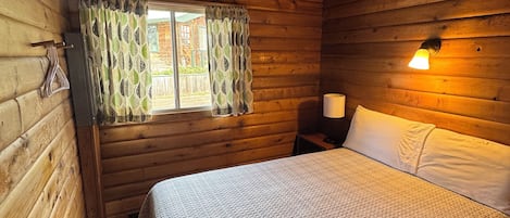 A wooden cabin room with a bed, a window with curtains, and a wall-mounted lamp.