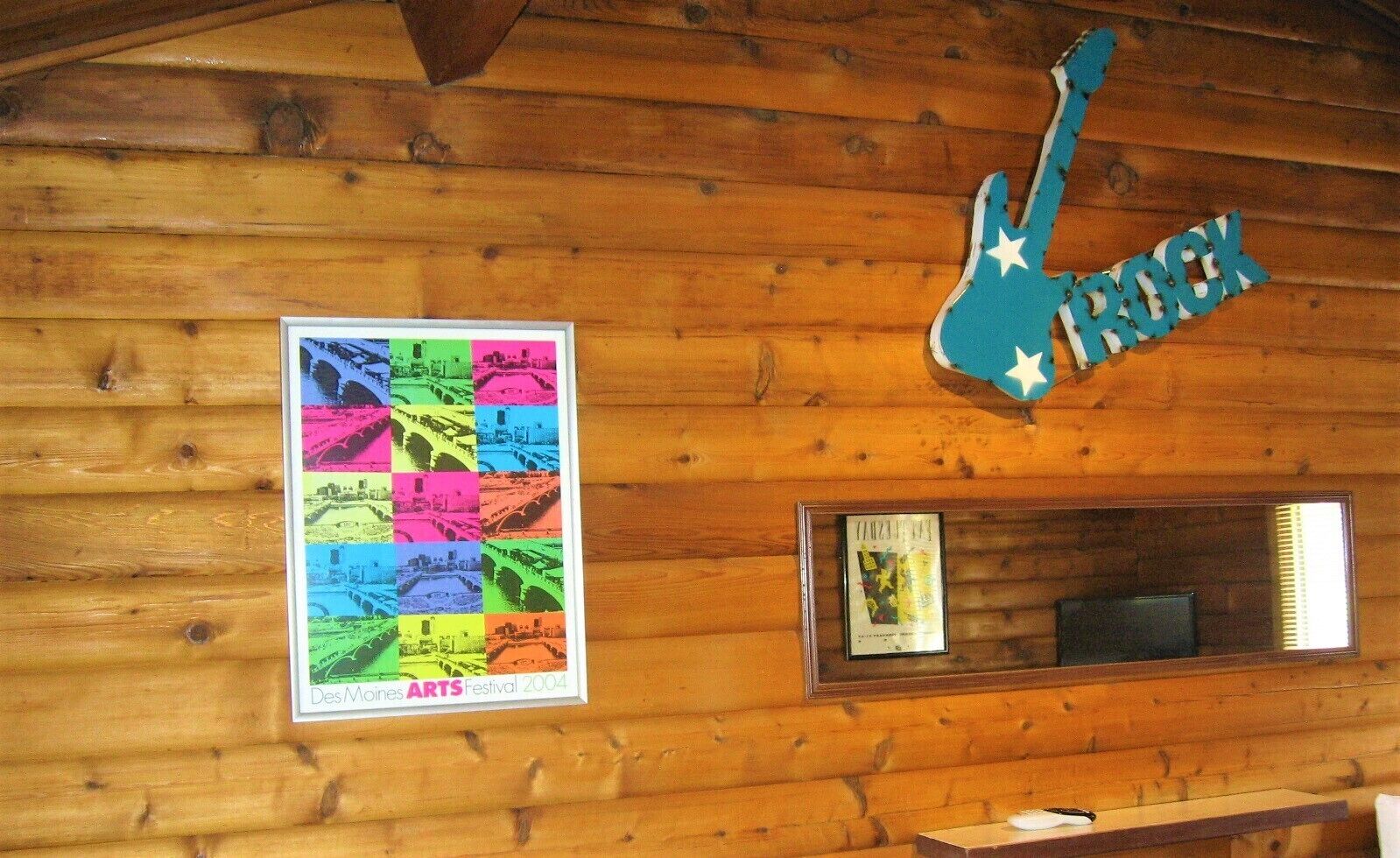 En trævæg med et indrammet plakat fra Des Moines Arts Festival 2004, et indrammet certifikat og et blåt guitarskilt med stjerner.