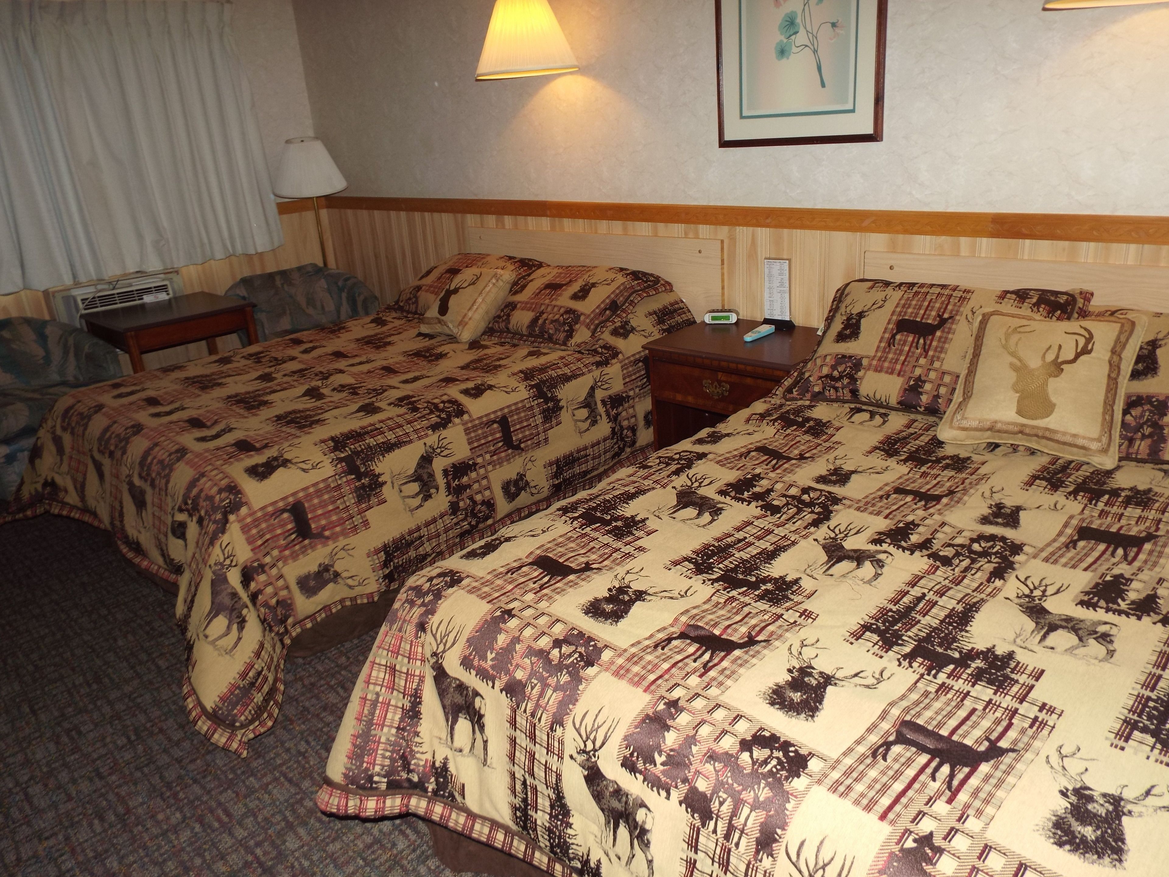 A hotel room with two beds, each with a patterned blanket featuring animal prints. There is a bedside table with a lamp, a wall-mounted clock, and a framed picture above the bed.