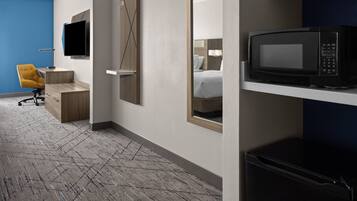 A hotel room with a bed, a TV mounted on the wall, a microwave on a shelf, and a desk with a chair.