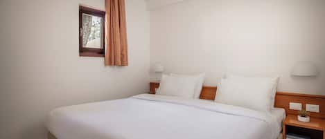 A neatly made bed with white linens, a wooden headboard, and a small bedside table with a lamp and a plant.