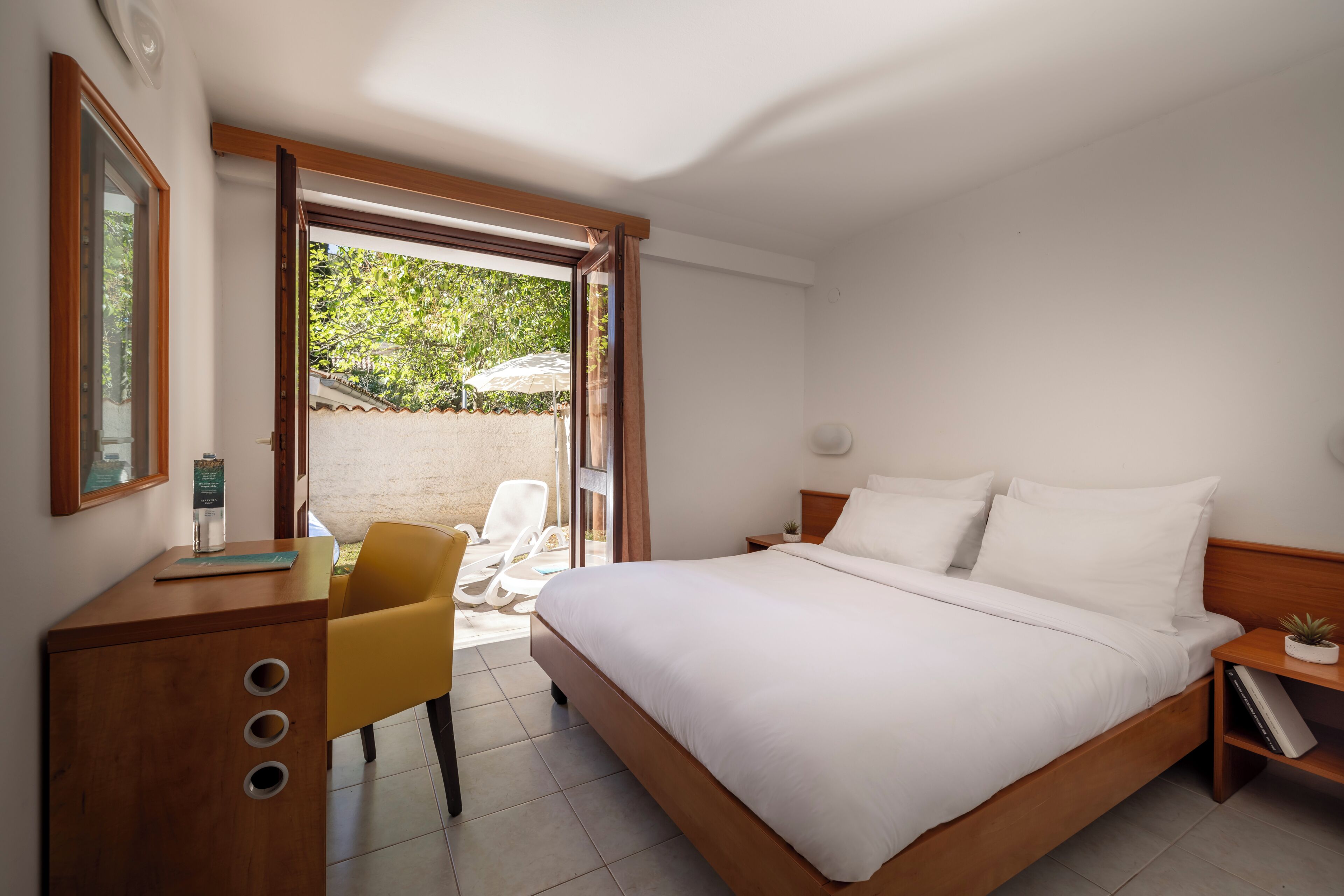 Un dormitorio con una cama grande, un escritorio de madera, una silla amarilla y un ventanal corredero que da a un espacio exterior.