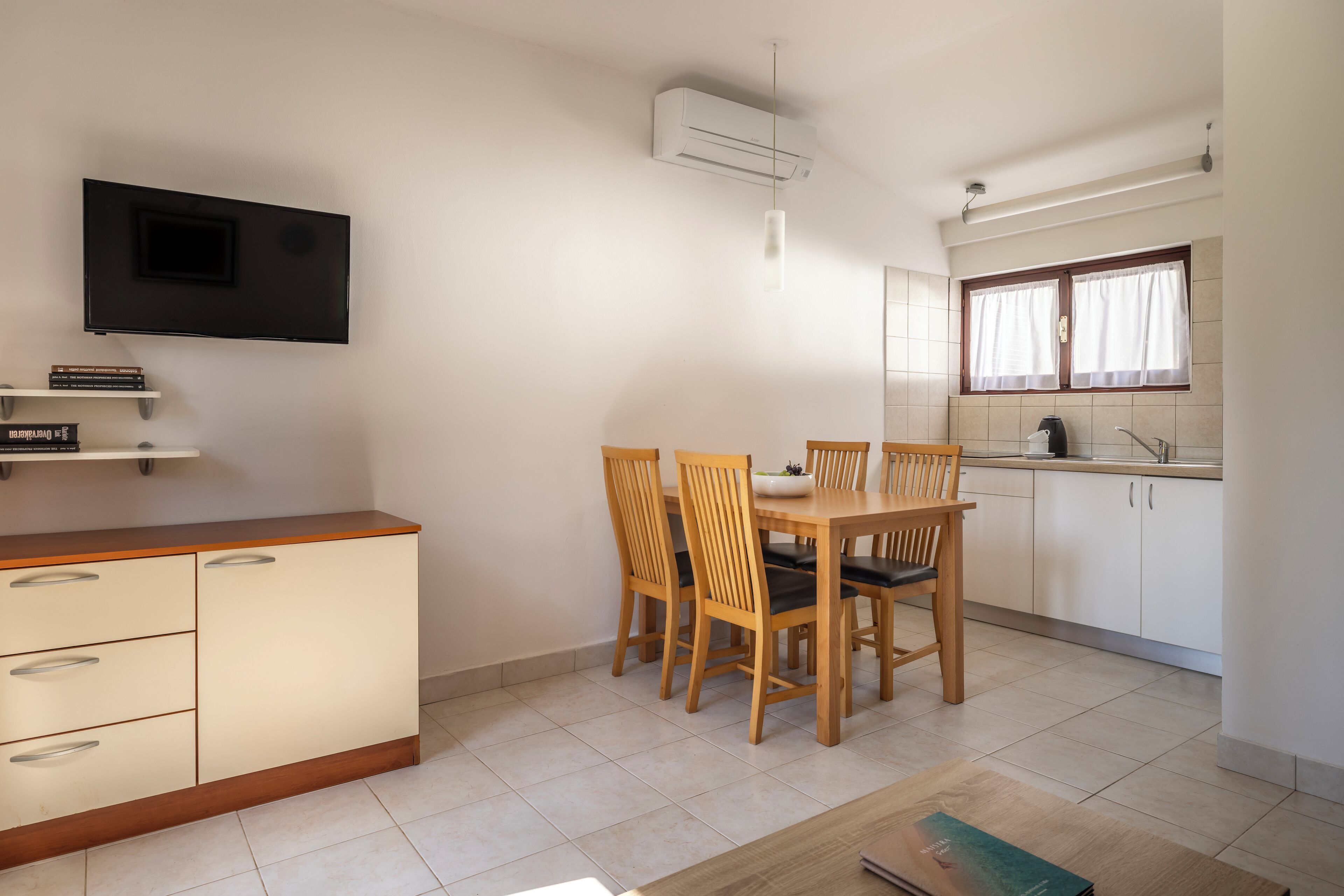 Una cocina funcional con mesa, sillas, televisor montado en la pared y ventana con cortinas.