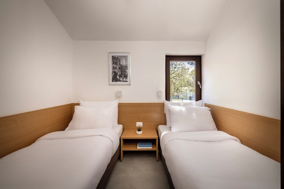 Habitación doble con dos camas, una mesita de noche, una pequeña mesa, un cuadro enmarco y una ventana.