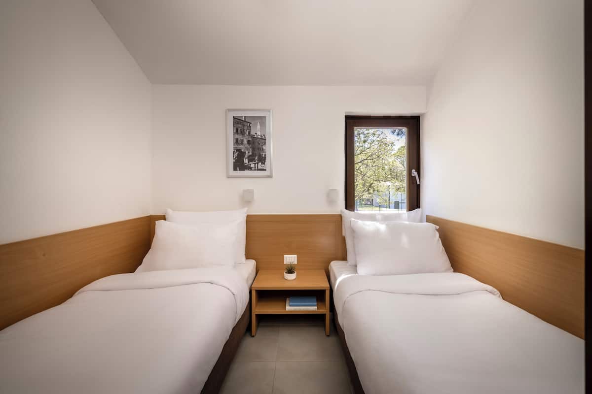 A twin room with two beds, a nightstand, a small table, a framed picture, and a window.