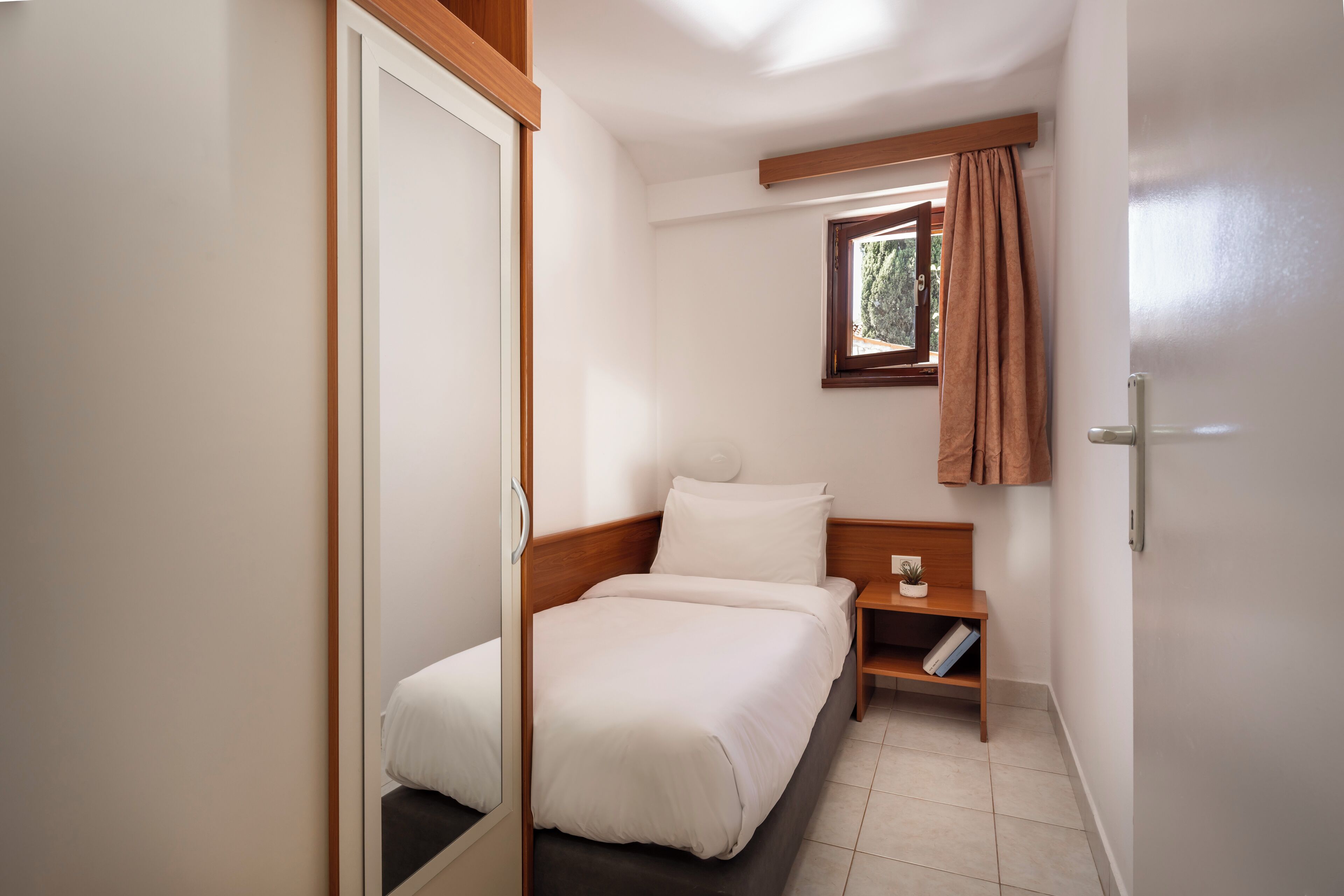A small, neatly arranged bedroom with a single bed, a bedside table, and a window with curtains.