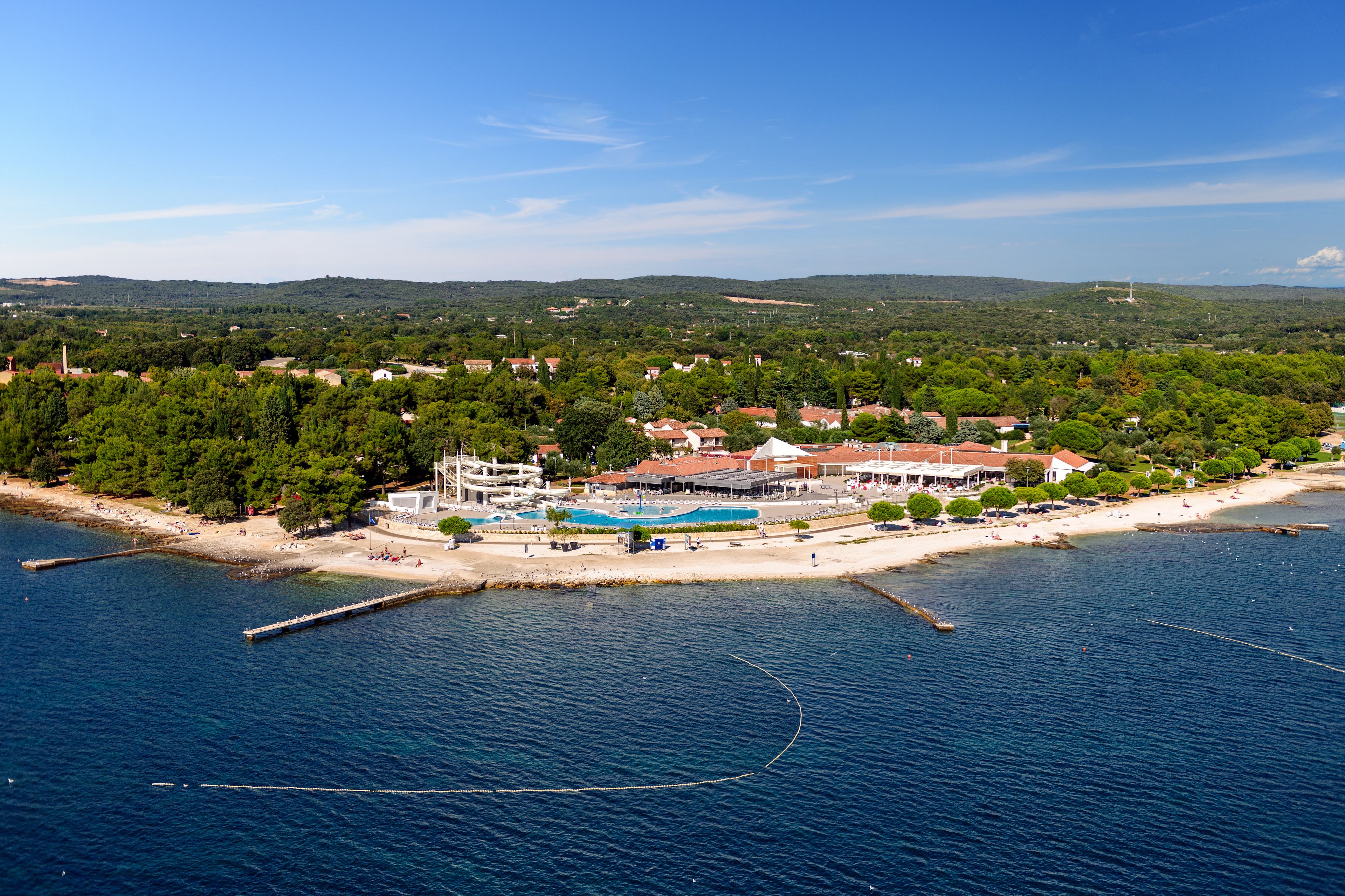 An aerial view of a coastal resort with a swimming pool, beach, and surrounding greenery.