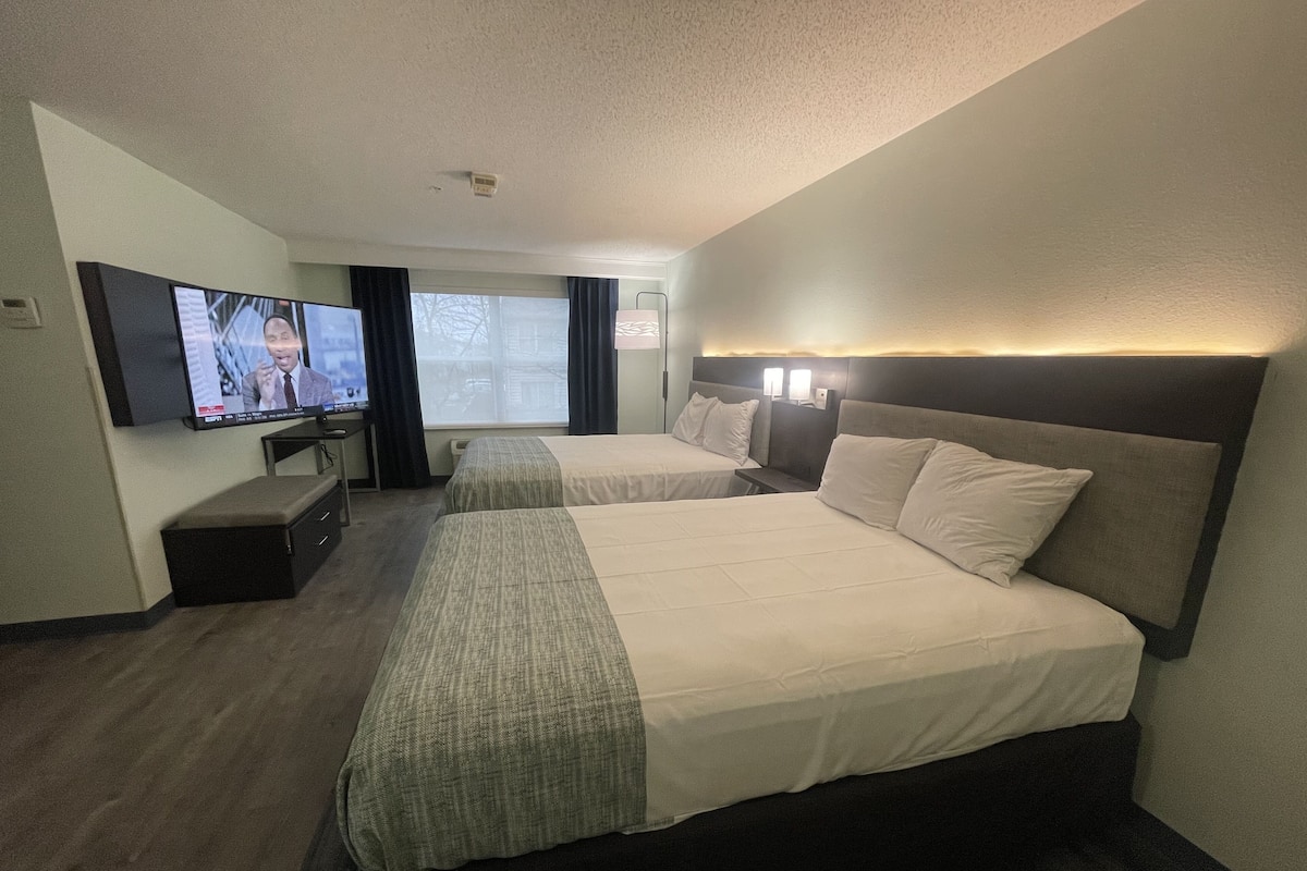 Une chambre d’hôtel avec deux lits, un téléviseur fixé au mur et une fenêtre avec des rideaux.